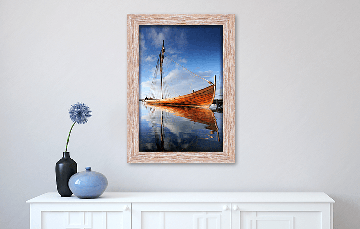 Decorative MDF Frame in red oak decor, with picture of a boat, placed in interior with white cupboard and vases. Picture frame producer Debex Suisse AG.