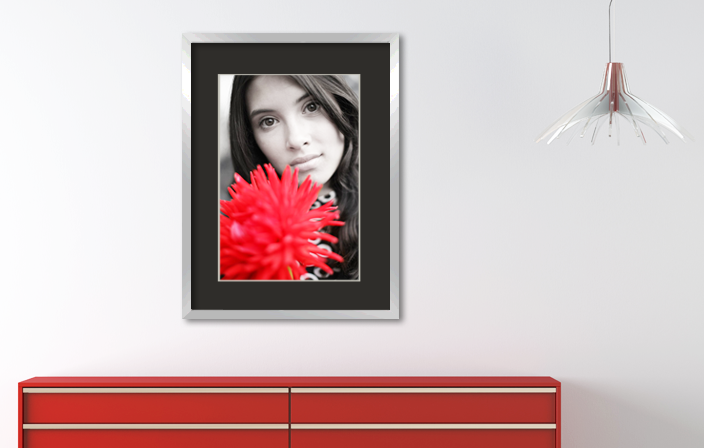 Aluminium picture frame with black mat board and a poster of a girl with red flower, placed in interior with red cupboard and a lamp. Picture frame producer Debex Suisse AG.